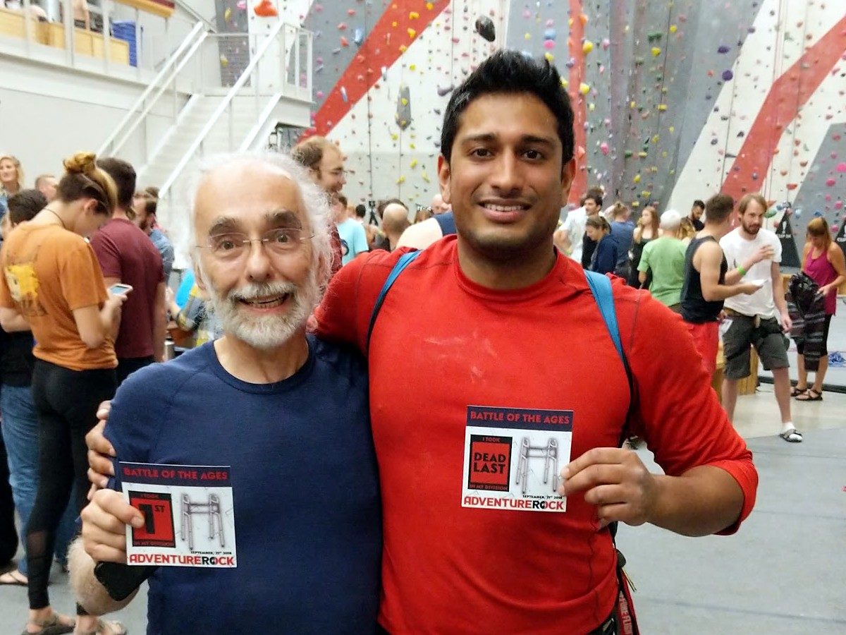 Two climbers at Adventure Rock’s Battle of the Ages competition, arms around each other. One holds a card reading “I took 1st in my division,” the other a card reading “I took dead last in my division.”