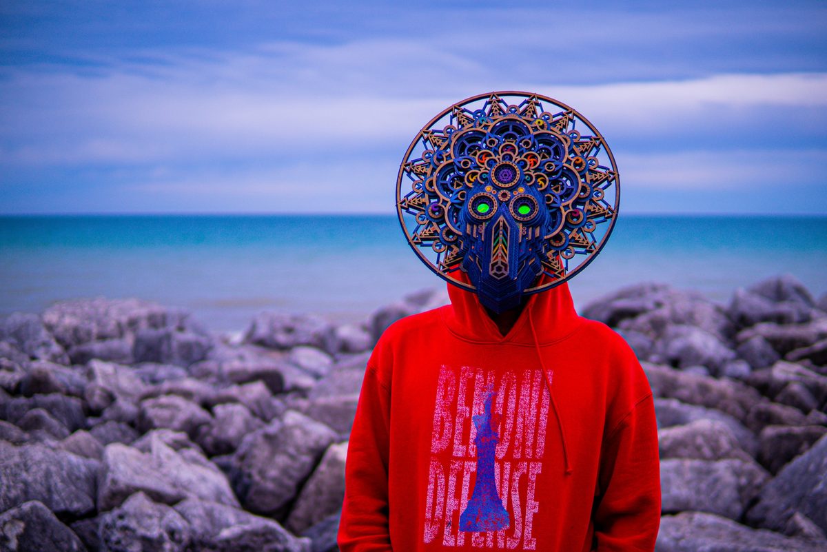 A person in a red hoodie printed with “Benoni Defense” and a chess king, wearing an ornate layered mandala mask with glowing green eyes, standing on boulders in front of a lake.