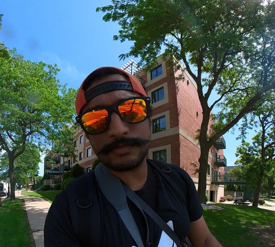 A wide-angle selfie of a man in a backwards cap and orange mirrored sunglasses, on a tree-lined sidewalk in summer with a brick apartment building behind.