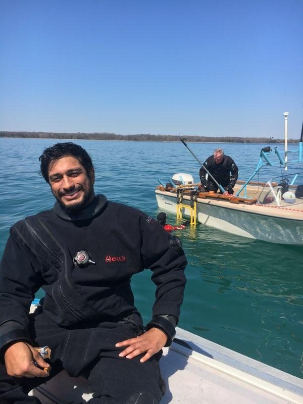 A diver in a black drysuit sitting on a boat, smiling, with open water and a second dive boat behind.