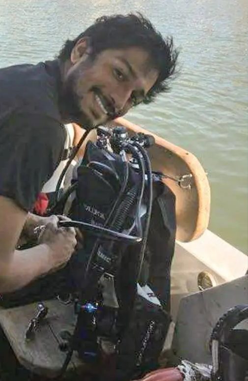 A diver on a boat in a harbor, grinning while assembling a regulator and wing on a tank.