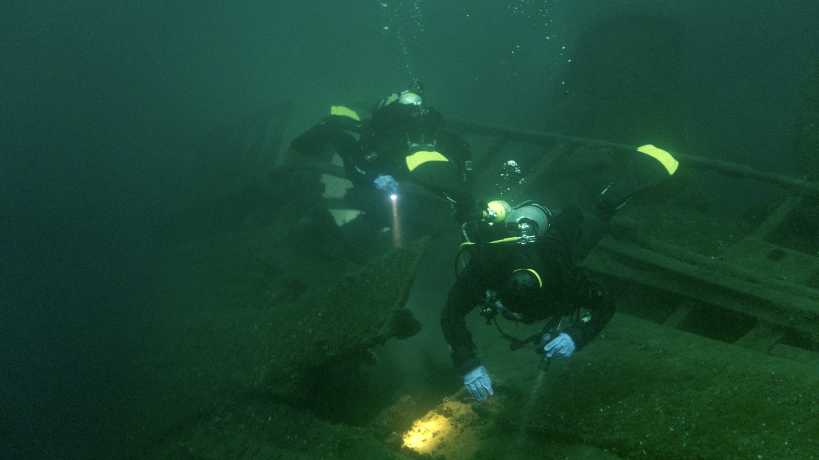 Two technical divers in drysuits, one sweeping a light across a wooden shipwreck in green water.